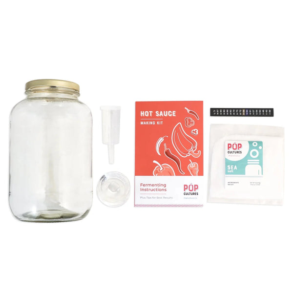 Hot sauce making kit with glass jar, airlock, fermentation weight, sea salt, LCD thermometer strip, instructions, and packaging on a white background